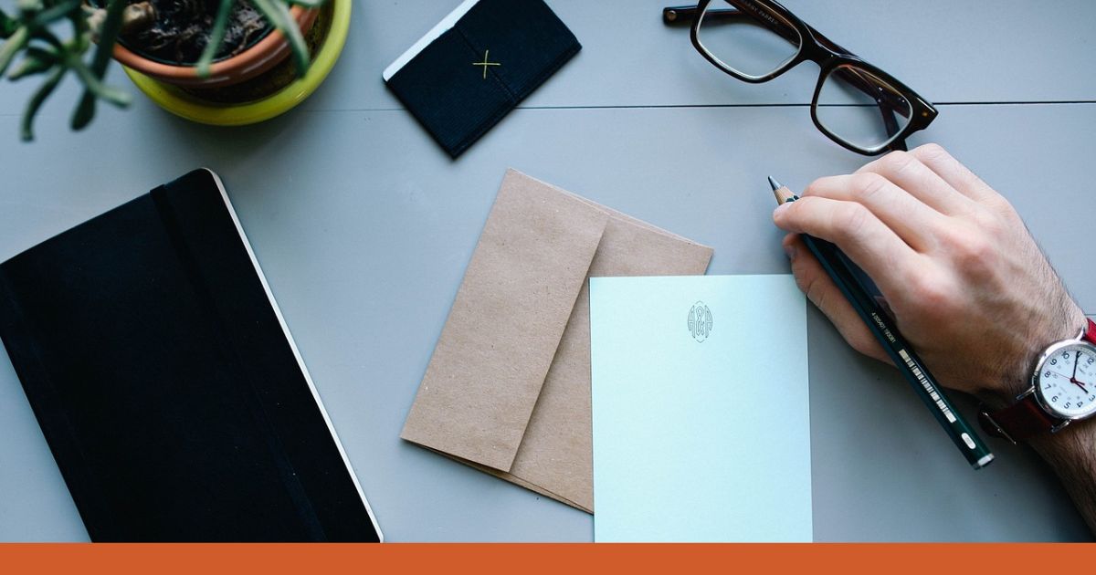 Auf einem Schreibtisch liegen Gegenstände aus dem Büro-Alltag: Ein Notizbuch, 2 Briefe, eine Hand die einen Stift hält, eine Brille und ein Etui.