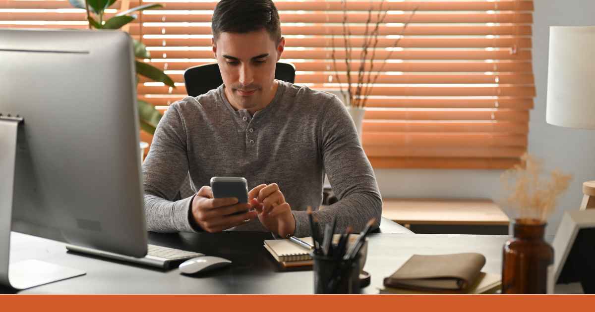 An einem Schreibtisch sitzt ein Mitarbeiter und blickt auf sein Smartphone. Er bereitet sich auf eine Telefonat zur Verhandlung mit einem Kunden vor.